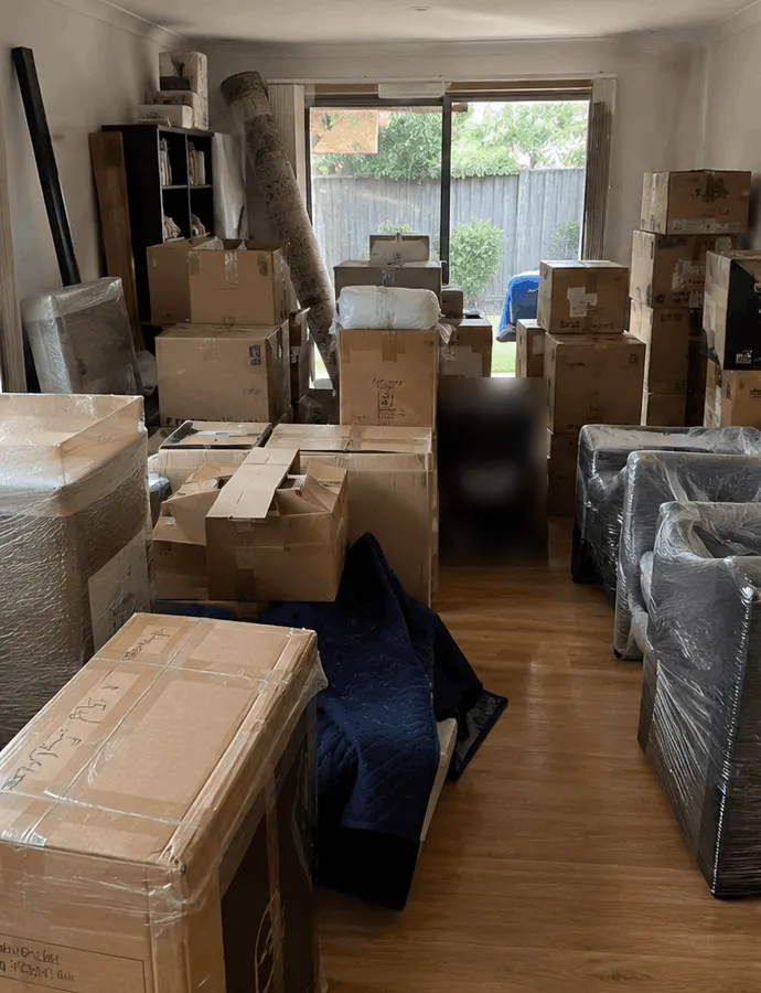 The same Melbourne lounge room on moving day: packed cartons stacked to the window and armchairs shrink-wrapped