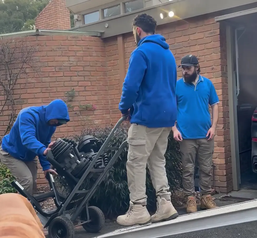 Three House Removalists Melbourne crew members in blue easing a heavy item down the truck ramp at a Melbourne brick home